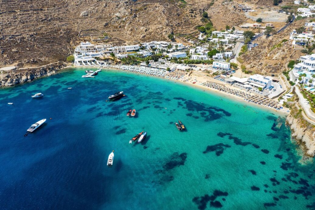 An aerial view of a beach in Mykonos, featuring clear blue waters, yachts, and sunbathers on the sandy shore.