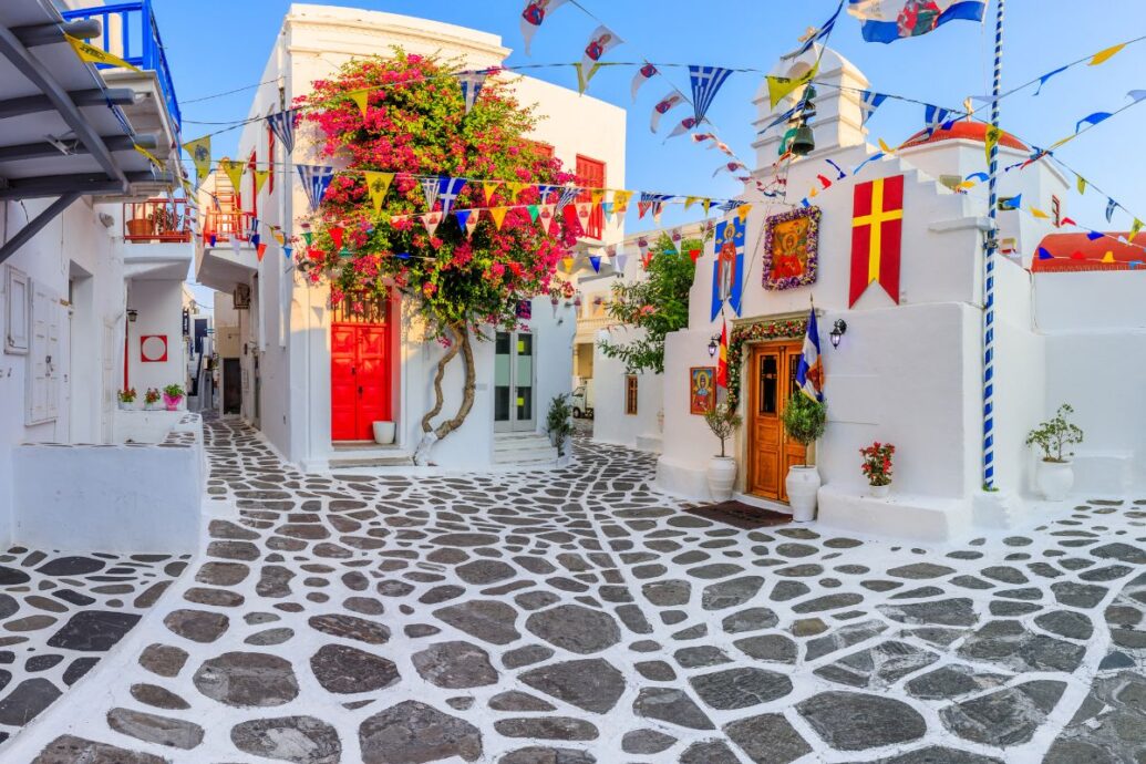 A charming street in Mykonos adorned with colorful flags, vibrant bougainvillea, and traditional white buildings, including a quaint church.