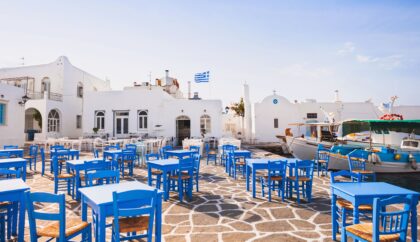 The image shows a picturesque outdoor café in Naoussa, Paros, with bright blue chairs and tables arranged on a stone-paved courtyard. Traditional white-washed buildings and a small church are in the background, along with a Greek flag fluttering above, capturing the charming and authentic atmosphere of the Cycladic island.