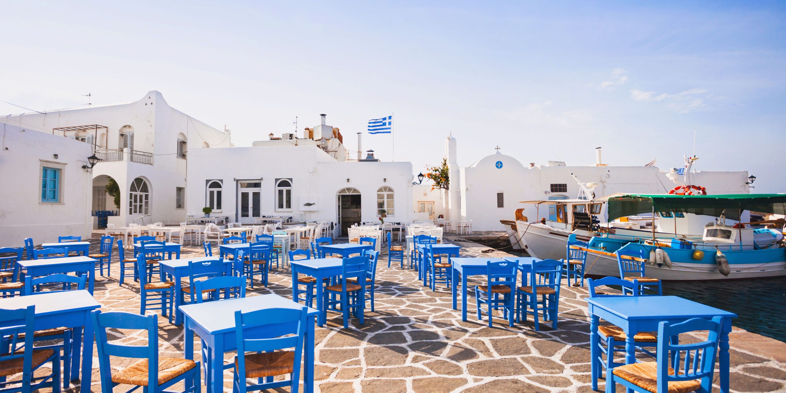The image shows a picturesque outdoor café in Naoussa, Paros, with bright blue chairs and tables arranged on a stone-paved courtyard. Traditional white-washed buildings and a small church are in the background, along with a Greek flag fluttering above, capturing the charming and authentic atmosphere of the Cycladic island.