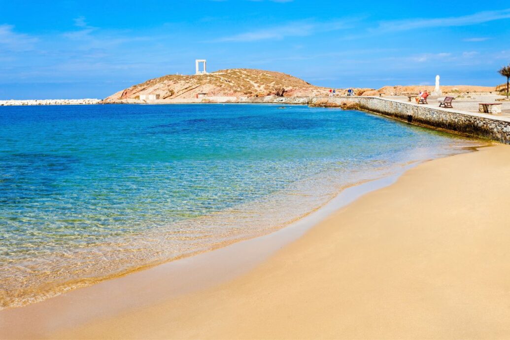 A tranquil view of Agios Georgios Beach in Naxos, featuring golden sands leading to clear, calm waters, with the iconic Portara visible on a hill in the background under a bright blue sky.