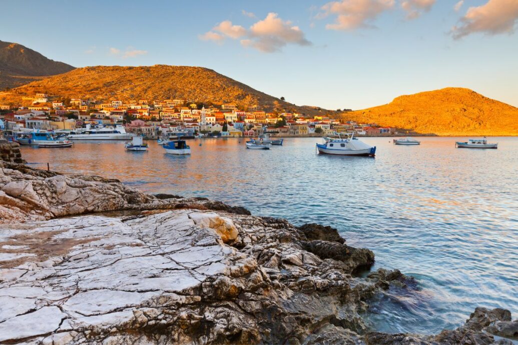A picturesque view of Halki Village in Naxos, with colorful houses nestled against sunlit hills, small boats floating in the calm waters of the harbor, and rocky shores in the foreground during a warm, golden sunset.