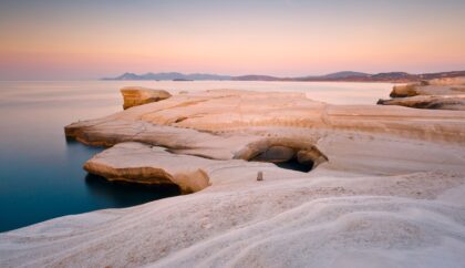The image shows the smooth, white rock formations of Sarakiniko Beach in Milos, Greece, gently sloping into calm, blue waters. The soft pastel colors of the sunset create a serene atmosphere, with distant mountains visible on the horizon. The unique, almost lunar landscape is bathed in warm, golden light.