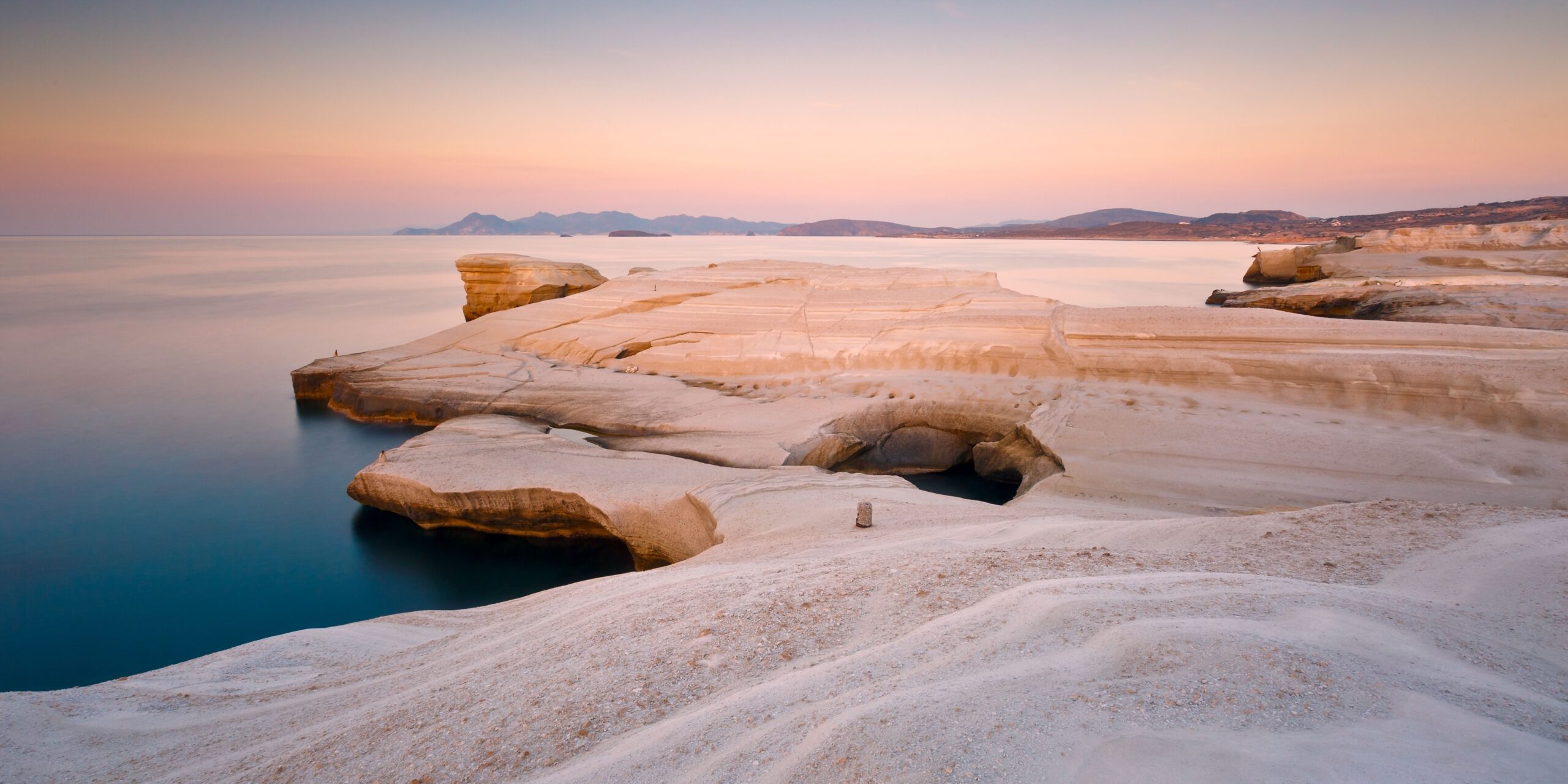 The image shows the smooth, white rock formations of Sarakiniko Beach in Milos, Greece, gently sloping into calm, blue waters. The soft pastel colors of the sunset create a serene atmosphere, with distant mountains visible on the horizon. The unique, almost lunar landscape is bathed in warm, golden light.