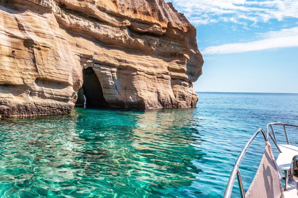 The image shows a rocky cliff formation with a sea cave along the coast of Milos, Greece. The clear turquoise water shimmers under the sunlight, and part of a boat is visible in the foreground, suggesting an exploration of the natural beauty of the island. The rock's layers and textures create a striking contrast against the calm sea.