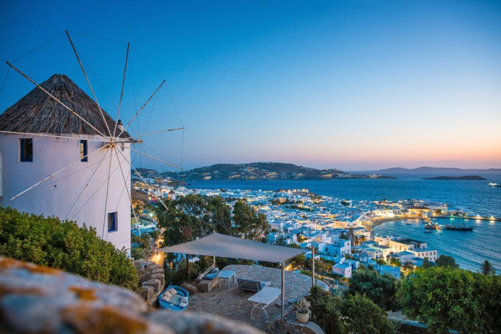 A stunning view of Mykonos Town at dusk, featuring a traditional windmill overlooking the whitewashed buildings, the bustling harbor, and the surrounding sea under a deepening blue sky.