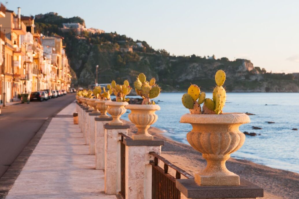 A scenic coastal promenade in Naxos Chora, lined with potted cacti and traditional buildings, overlooking the calm sea with a mountainous landscape in the background during the golden hour.