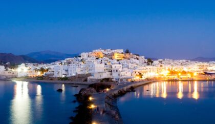 A serene evening view of Naxos Town, showcasing its charming whitewashed buildings illuminated by warm lights reflecting on the calm waters of the harbor, set against a backdrop of rolling hills under a deep blue sky.