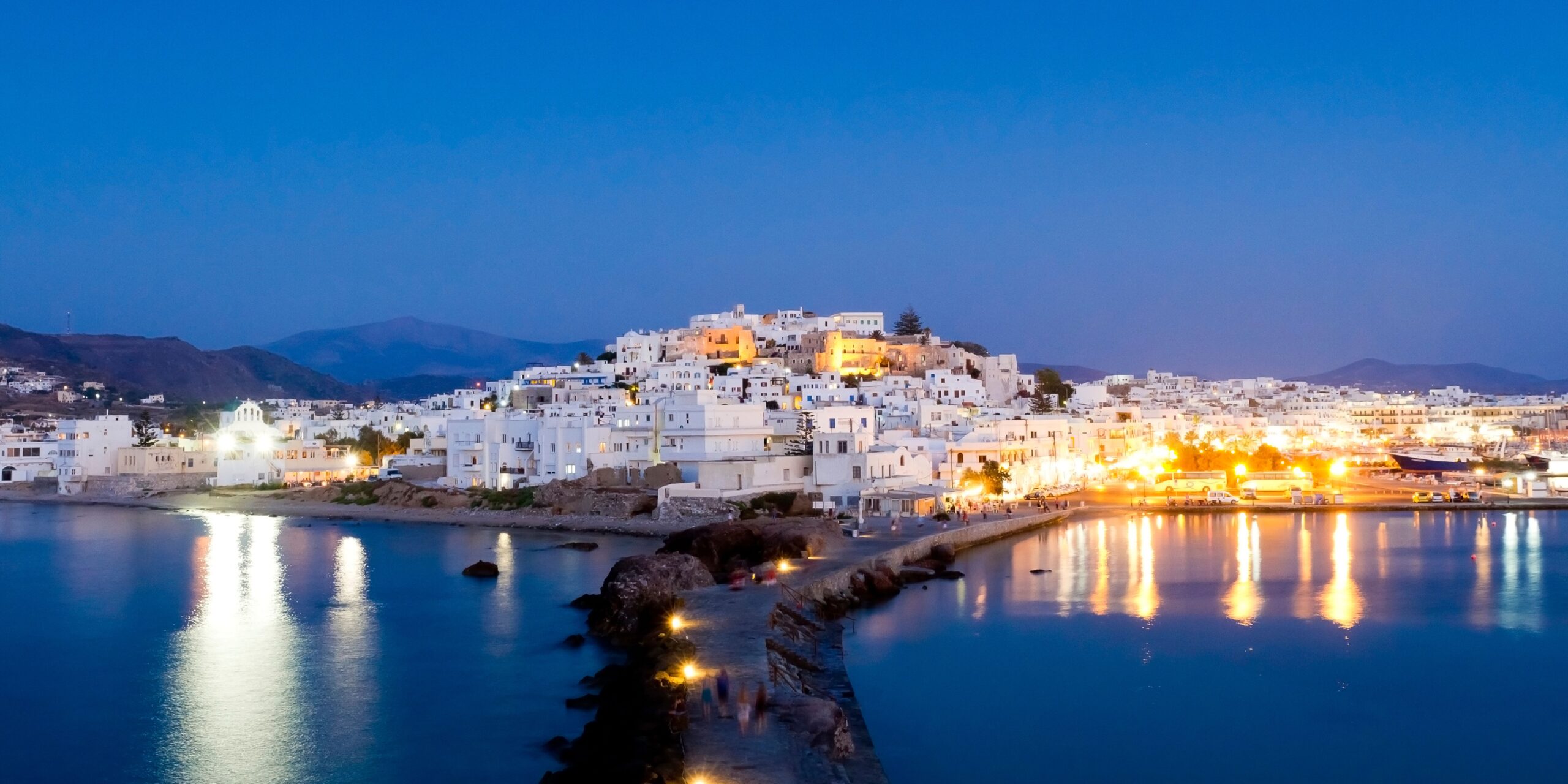 A serene evening view of Naxos Town, showcasing its charming whitewashed buildings illuminated by warm lights reflecting on the calm waters of the harbor, set against a backdrop of rolling hills under a deep blue sky.