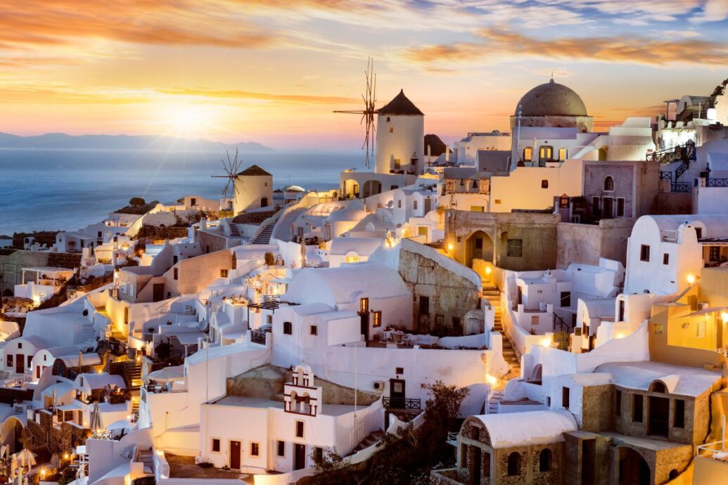 A stunning view of Oia, Santorini at sunset, featuring whitewashed buildings, windmills, and a soft golden glow as the lights come on, with the Aegean Sea in the background.