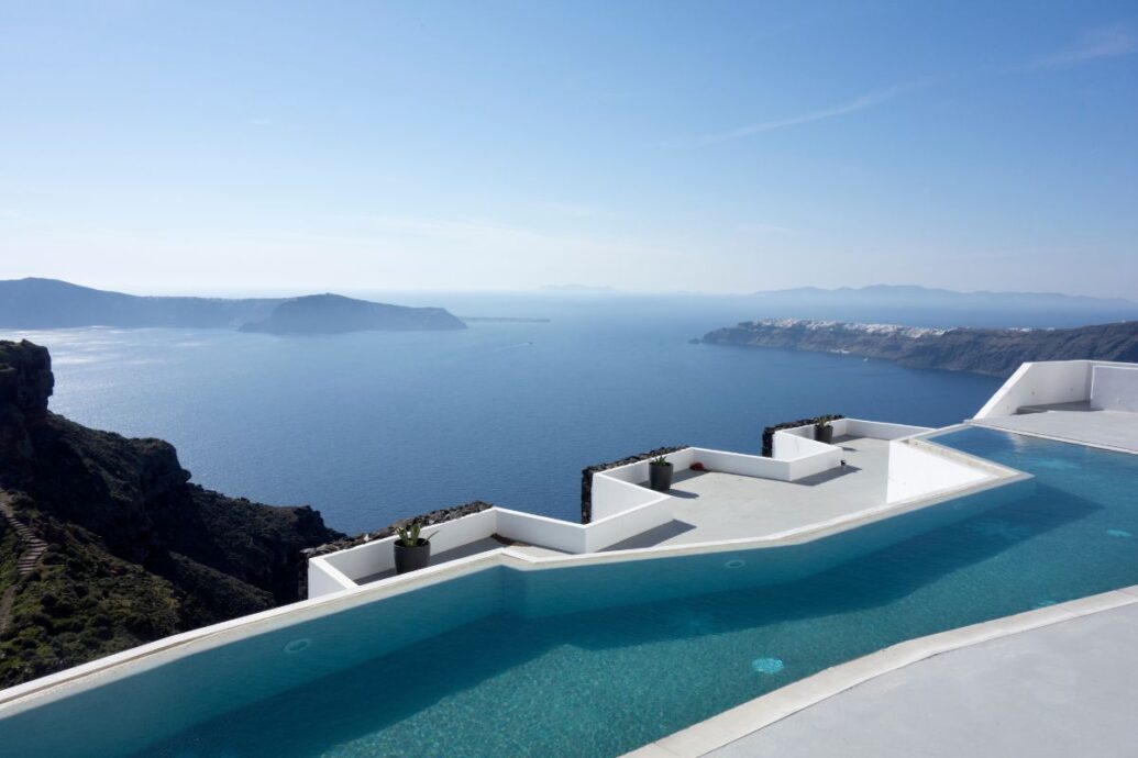 A serene view from a luxury Santorini hotel overlooking an infinity pool that blends into the vast blue sea and distant islands, with clear skies enhancing the peaceful atmosphere.