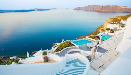 A picturesque Santorini hotel with whitewashed terraces and a staircase leading down to an infinity pool overlooking the calm blue sea and distant islands, creating a tranquil and luxurious atmosphere.