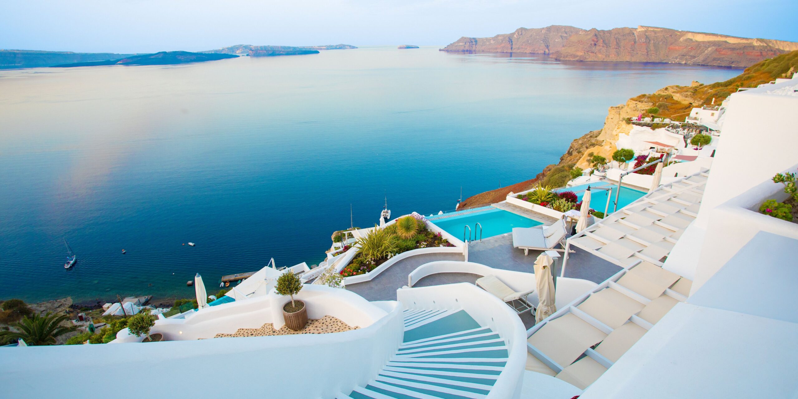 A picturesque Santorini hotel with whitewashed terraces and a staircase leading down to an infinity pool overlooking the calm blue sea and distant islands, creating a tranquil and luxurious atmosphere.