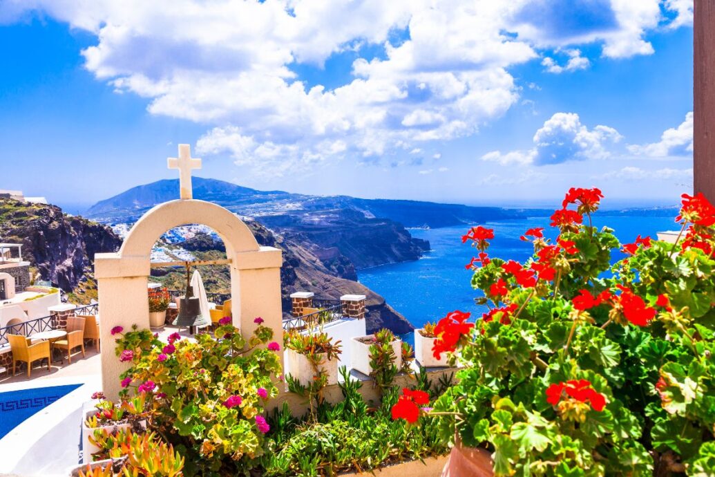 A vibrant scene of blooming flowers in Santorini during spring, with a white archway topped by a cross overlooking the deep blue sea and rocky cliffs under a bright, clear sky.