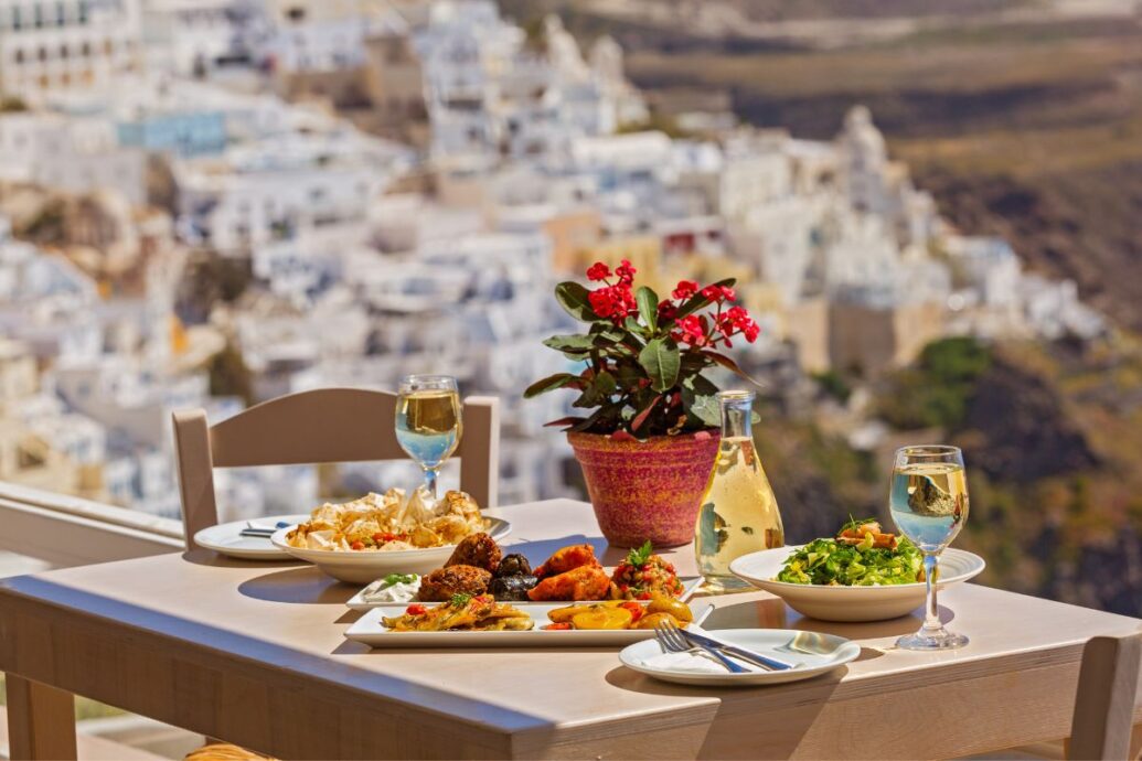 A table set with traditional Greek dishes, wine glasses, and a carafe of white wine, with a vibrant flower pot in the center, overlooking the scenic whitewashed buildings of Santorini in the background.