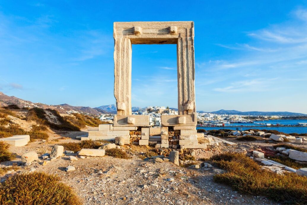 The image showcases the ancient marble gateway known as the Portara, the iconic entrance to the unfinished Temple of Apollo on Naxos Island, with the town and sea visible in the background under a bright blue sky.