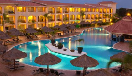 A serene view of a luxurious all-inclusive resort in Corfu, featuring a large, illuminated pool surrounded by palm trees and thatched sun loungers, with balconies overlooking the scene.
