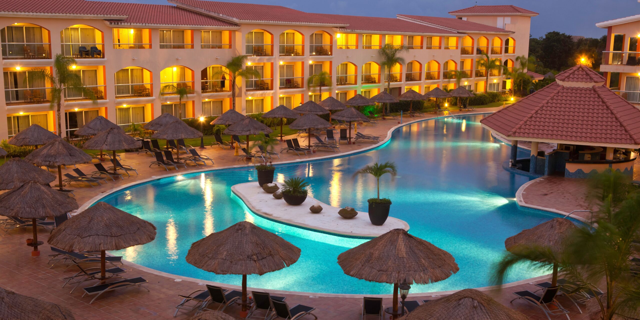 A serene view of a luxurious all-inclusive resort in Corfu, featuring a large, illuminated pool surrounded by palm trees and thatched sun loungers, with balconies overlooking the scene.