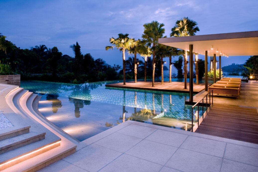 A luxurious hotel pool in Corfu, illuminated in the evening, surrounded by palm trees and sleek lounge areas, with tranquil water reflecting the soft lighting and lush greenery.
