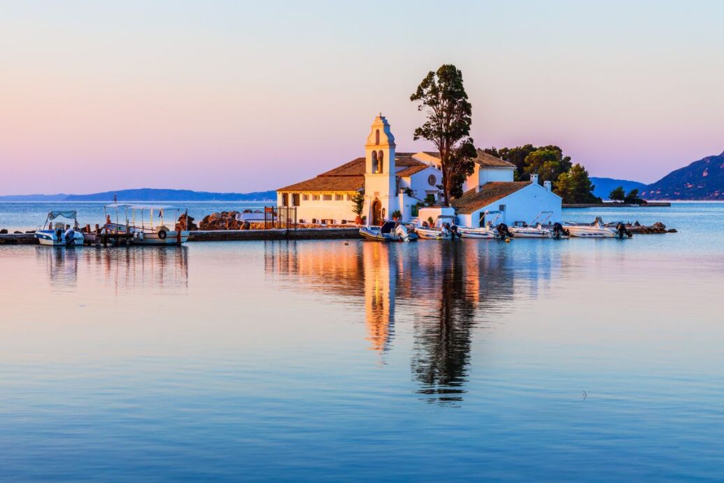 A tranquil scene in Corfu showcasing a small whitewashed church by the water, with boats docked along the pier, bathed in soft sunset hues reflecting on the calm sea.