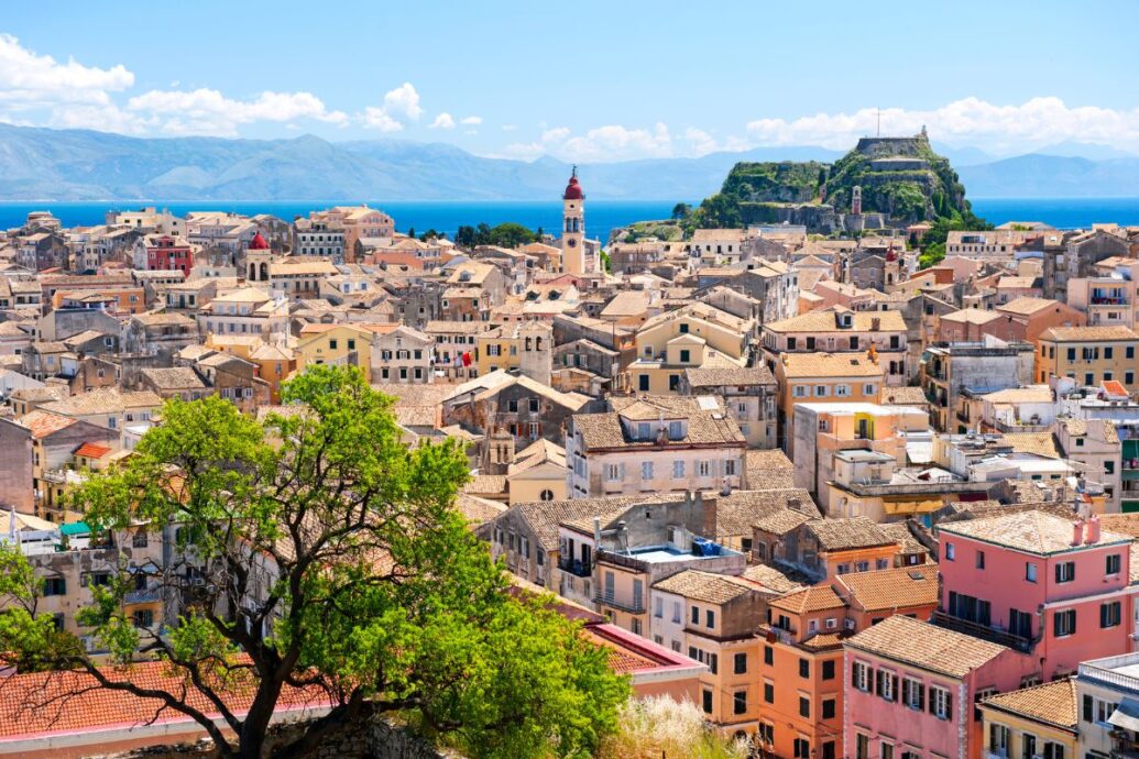 A scenic view of Corfu's Old Town, featuring a dense cluster of historic buildings with terracotta roofs, a bell tower rising in the center, and a fortress perched in the distance, all set against the backdrop of the sea and distant mountains.