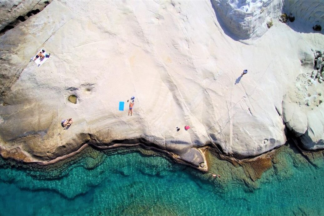 The image shows an aerial view of a beach in Milos, Greece, featuring smooth white rock formations that slope gently into clear turquoise water. A few people are sunbathing on the rocks, and one person is standing near a towel, enjoying the serene, natural setting. The contrast between the bright rock and the calm sea creates a striking, tranquil scene.