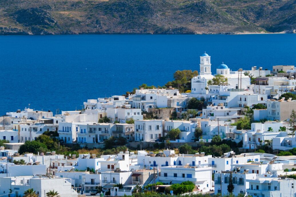 The image shows a coastal town in Milos, Greece, with whitewashed buildings and blue-domed churches overlooking the deep blue sea. The town is nestled against a backdrop of rugged hills, creating a picturesque contrast between the architecture and natural surroundings.