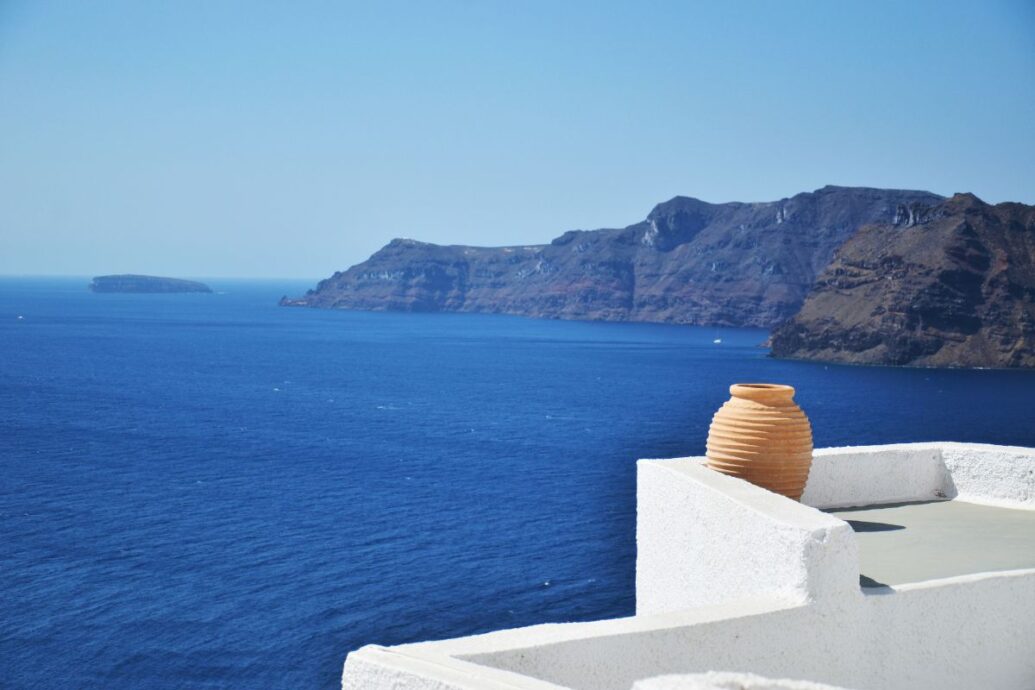 A simple Santorini scene with a terracotta pot resting on a whitewashed terrace, overlooking the deep blue Aegean Sea and rocky cliffs in the distance under a clear sky.