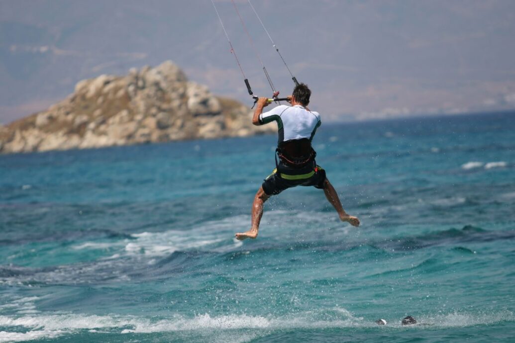 A kite surfer catching air above the turquoise waters of Naxos, with a rocky island in the background, showcasing the island's popular water sports activities.