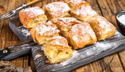 The image shows a serving of a golden-brown Greek pastry dusted with powdered sugar, cut into square portions and presented on a wooden board. The flaky layers of the pastry are visible, with a creamy filling inside. A fork and a small bowl of powdered sugar are placed nearby.