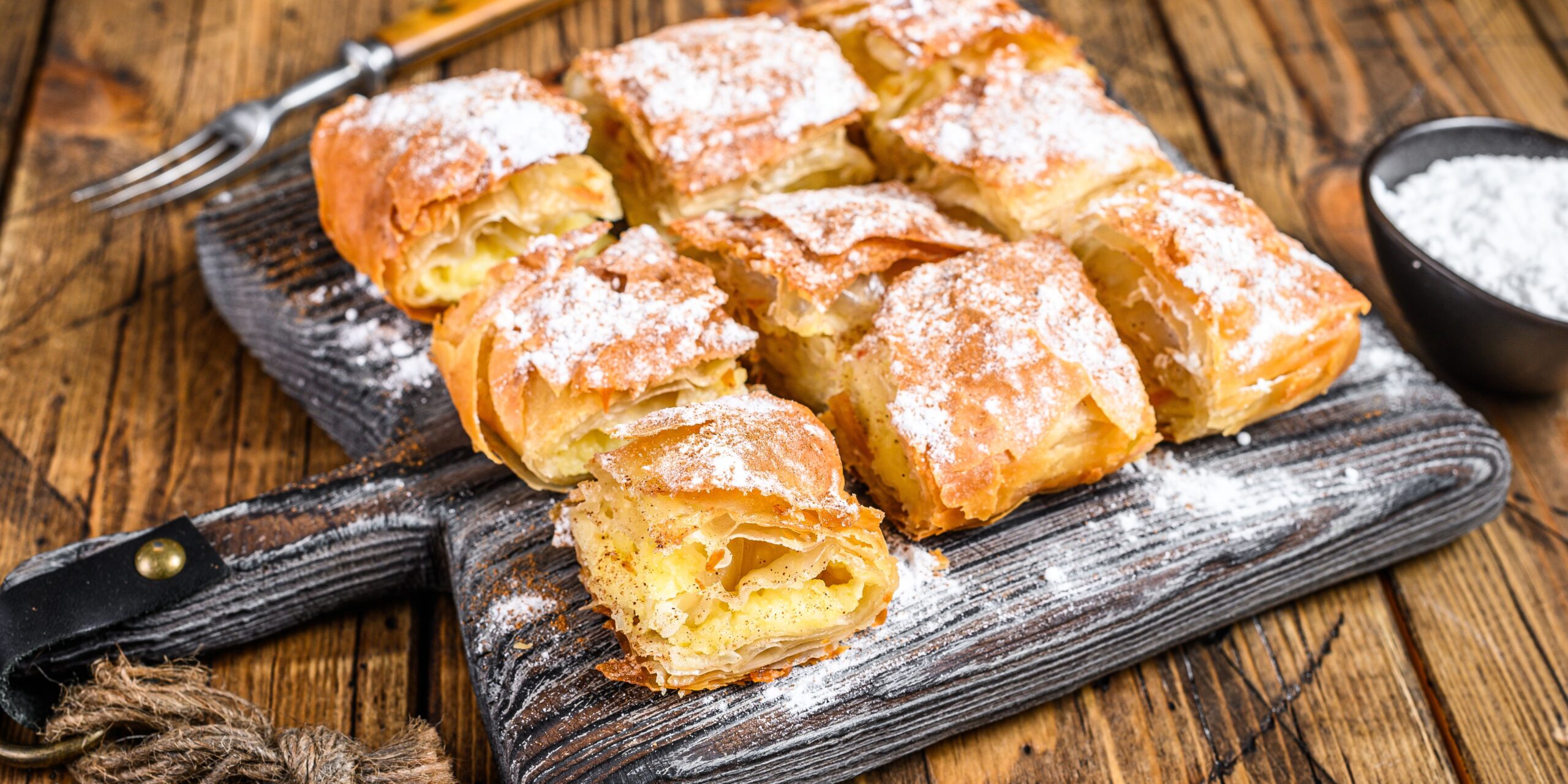 The image shows a serving of a golden-brown Greek pastry dusted with powdered sugar, cut into square portions and presented on a wooden board. The flaky layers of the pastry are visible, with a creamy filling inside. A fork and a small bowl of powdered sugar are placed nearby.