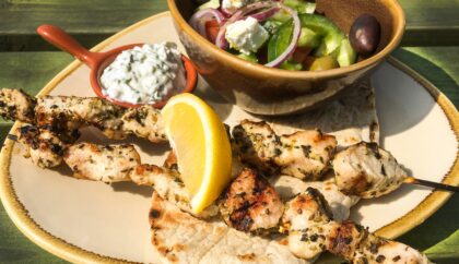 The image shows grilled chicken skewers served on pita bread, accompanied by a lemon wedge, a small bowl of tzatziki, and a Greek salad with feta cheese, olives, and vegetables.