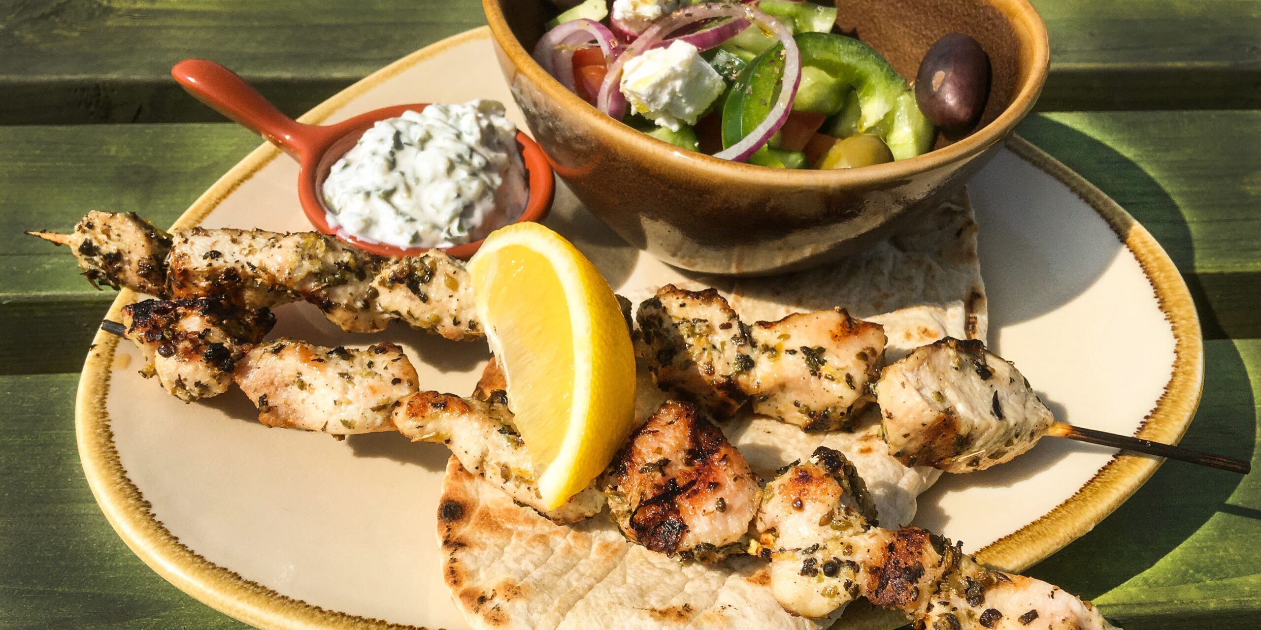 The image shows grilled chicken skewers served on pita bread, accompanied by a lemon wedge, a small bowl of tzatziki, and a Greek salad with feta cheese, olives, and vegetables.