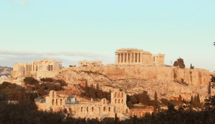 A view of the Acropolis in Athens, Greece, featuring ancient structures like the Parthenon, set against a warm, late afternoon sky.