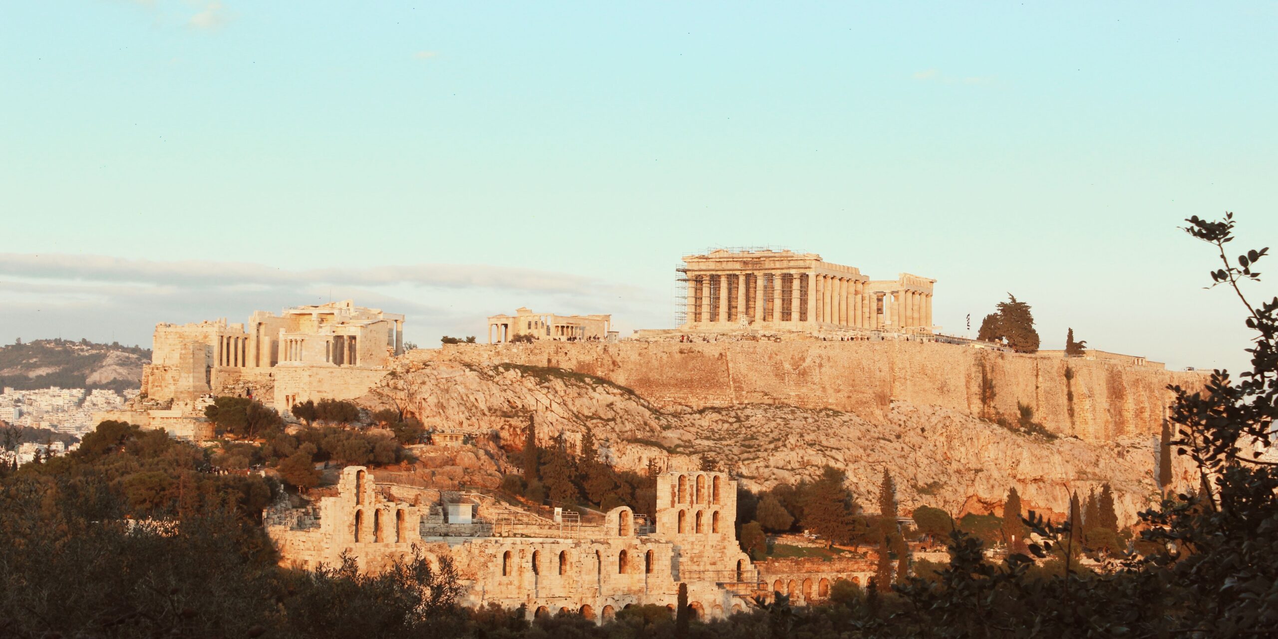 A view of the Acropolis in Athens, Greece, featuring ancient structures like the Parthenon, set against a warm, late afternoon sky.