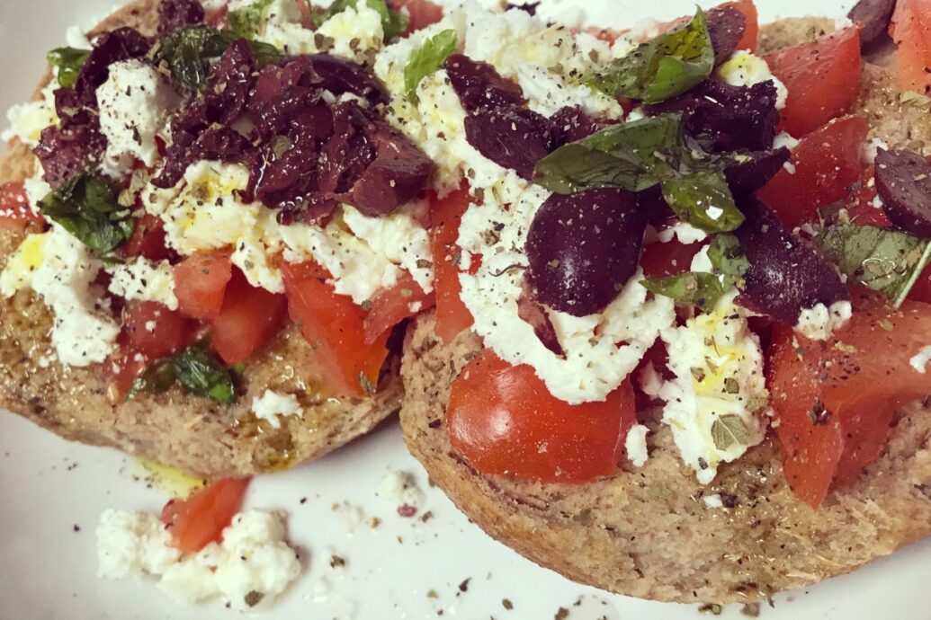 The image shows Cretan dakos, a traditional dish made with a barley rusk topped with chopped tomatoes, crumbled cheese, olives, fresh herbs, and a drizzle of olive oil.