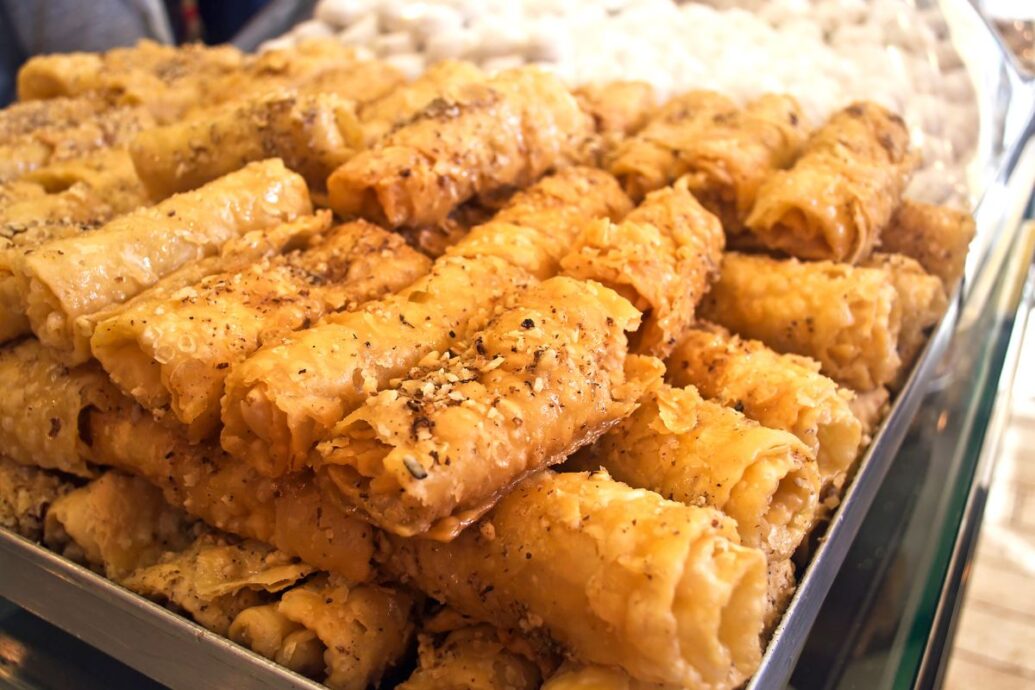The image shows a tray filled with Greek diples, a traditional dessert made from thin, fried dough rolls. The diples are golden and crispy, sprinkled with crushed nuts and drizzled with syrup.