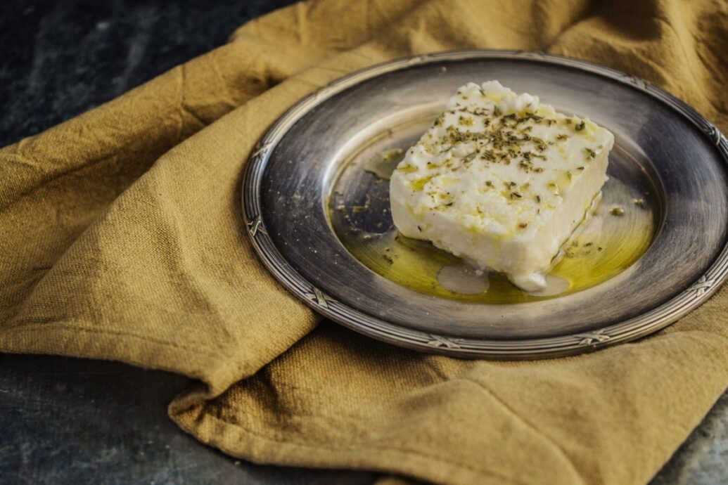 The image shows a slice of feta cheese drizzled with olive oil and sprinkled with herbs, served on a metal plate.