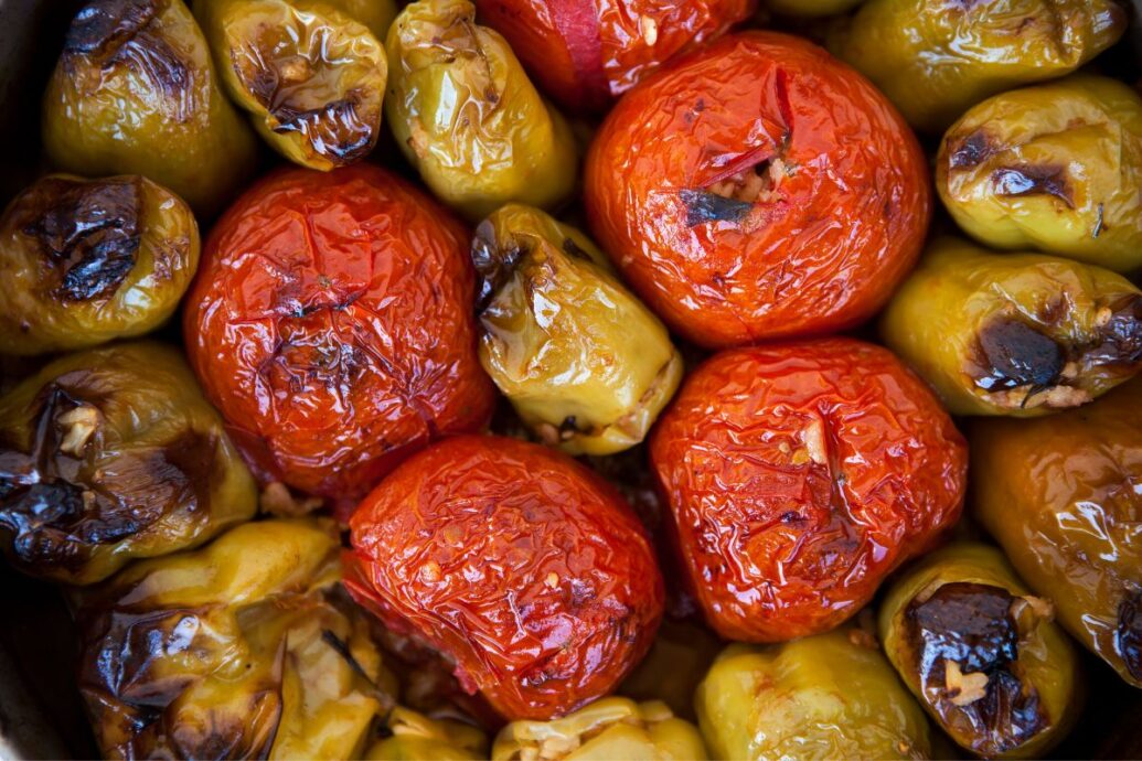 The image shows a tray of gemista, featuring roasted tomatoes and bell peppers stuffed with rice or other fillings. The vegetables are baked until soft, with a slightly charred and golden exterior.