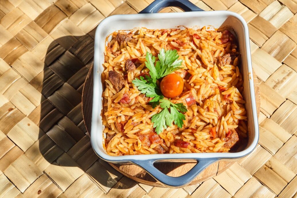 The image shows a dish of giouvetsi, a traditional Greek meal made with orzo pasta, cooked in a tomato-based sauce with meat, garnished with parsley and a cherry tomato.