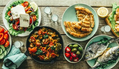 The image shows a variety of traditional Greek dishes, including grilled fish, fresh salads with feta, stuffed peppers, grilled meats, and phyllo pastries, all arranged on a wooden table.