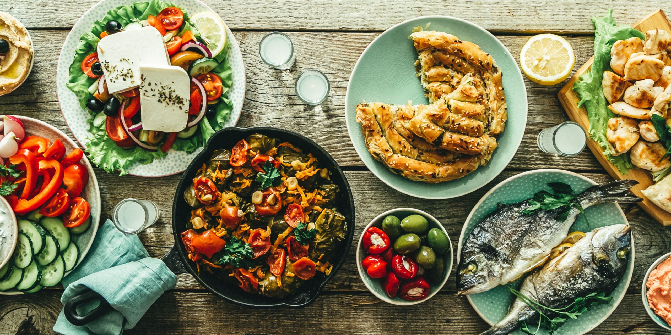 The image shows a variety of traditional Greek dishes, including grilled fish, fresh salads with feta, stuffed peppers, grilled meats, and phyllo pastries, all arranged on a wooden table.