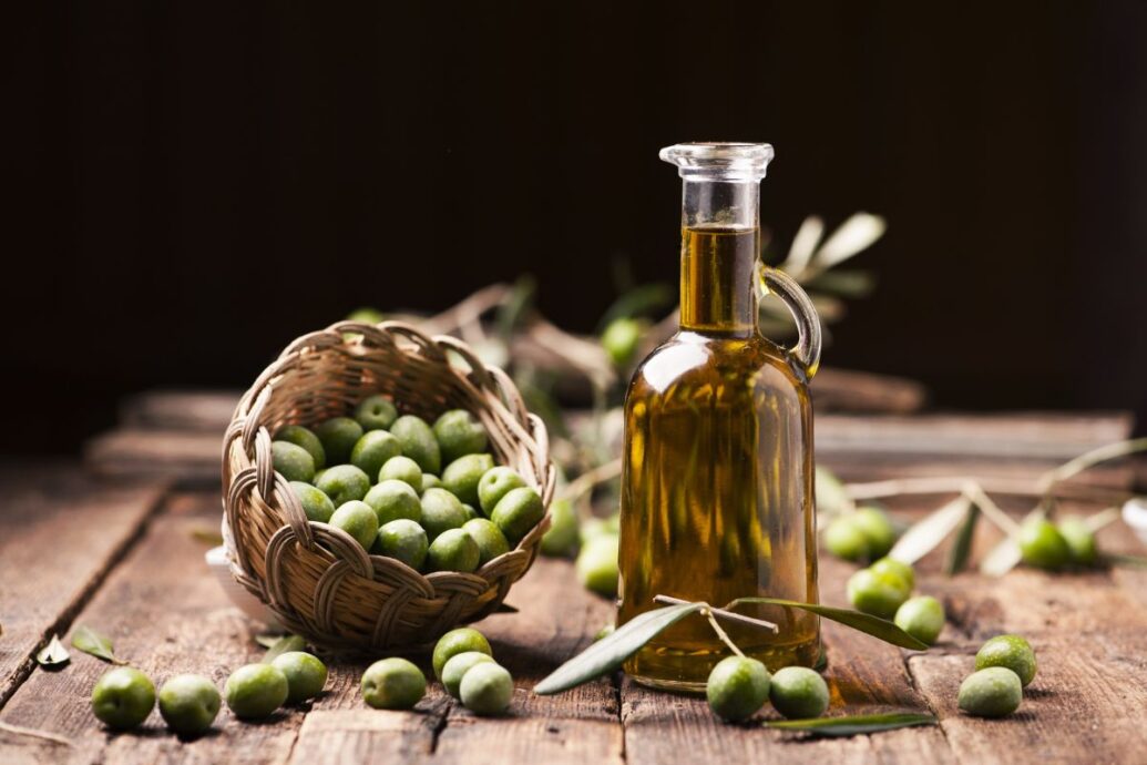 The image shows a bottle of olive oil next to a basket filled with green olives, with a few olives scattered around, set on a rustic wooden surface.