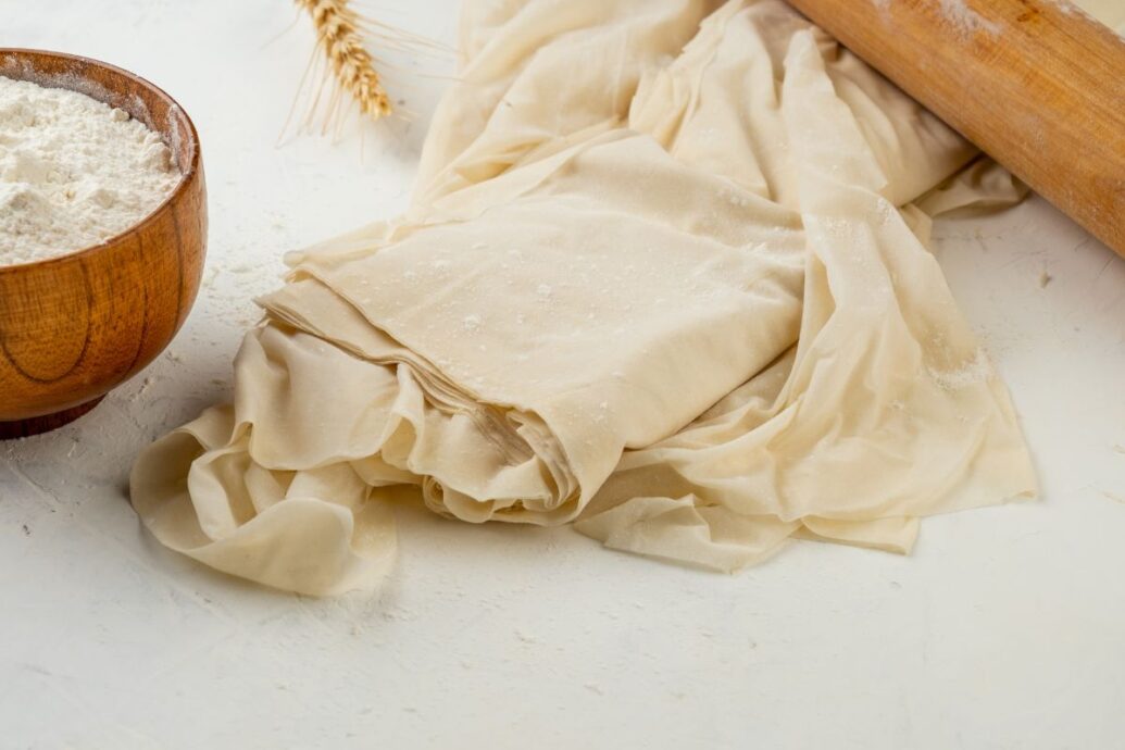 A sheet of thin Greek phyllo dough is spread out on a surface, lightly dusted with flour, alongside a wooden bowl filled with flour and a rolling pin.
