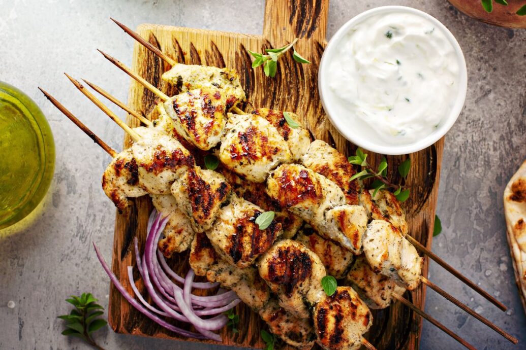 The image shows grilled chicken skewers served on a wooden board with a side of tzatziki sauce and some sliced red onions. Fresh herbs are scattered on top, enhancing the presentation.