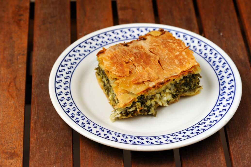 A single piece of Greek spanakopita, a spinach and cheese pie with layers of crisp, golden filo pastry, sits on a white plate with a blue decorative border. The dish highlights the savory filling and flaky crust.