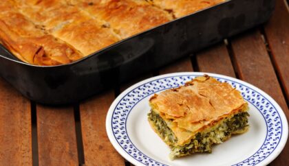 The image shows pieces of spanakopita, a traditional Greek savory pie made with flaky filo pastry and filled with spinach and cheese. The golden-brown, crisp layers are visible, giving the dish a mouthwatering appearance.