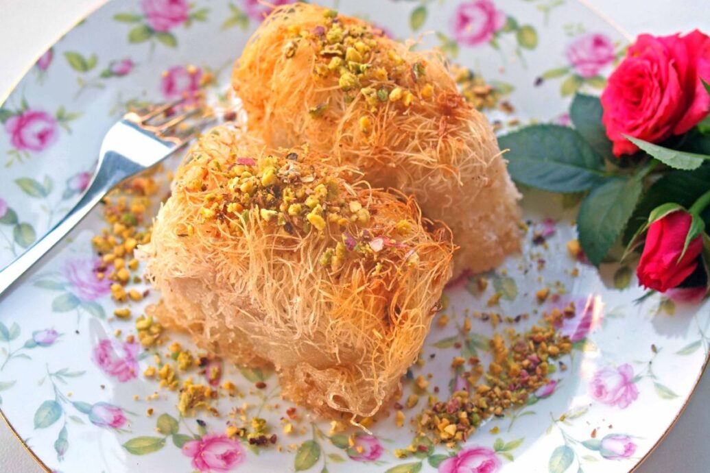 The image shows a plate of Greek kataifi, a dessert made from shredded phyllo dough, served in two portions and topped with crushed pistachios. The dessert sits on a floral-patterned plate, with a fork nearby and a red rose for decoration.