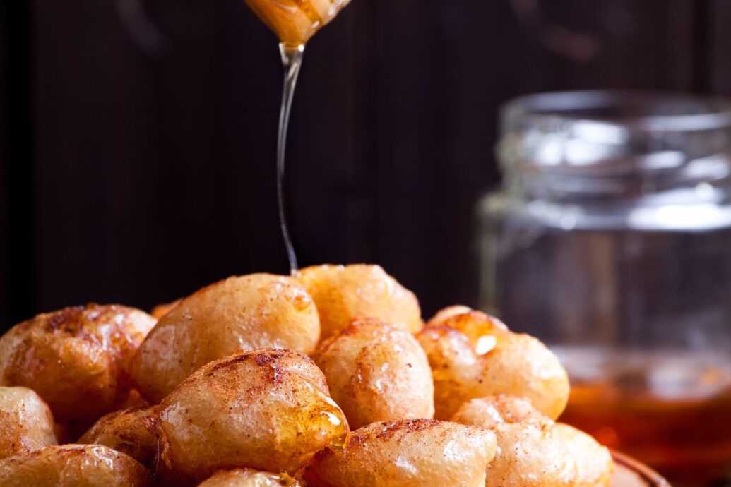 The image shows a plate of loukoumades, Greek fried dough balls, being drizzled with honey. The loukoumades are golden and crispy, coated with a light sprinkle of cinnamon.