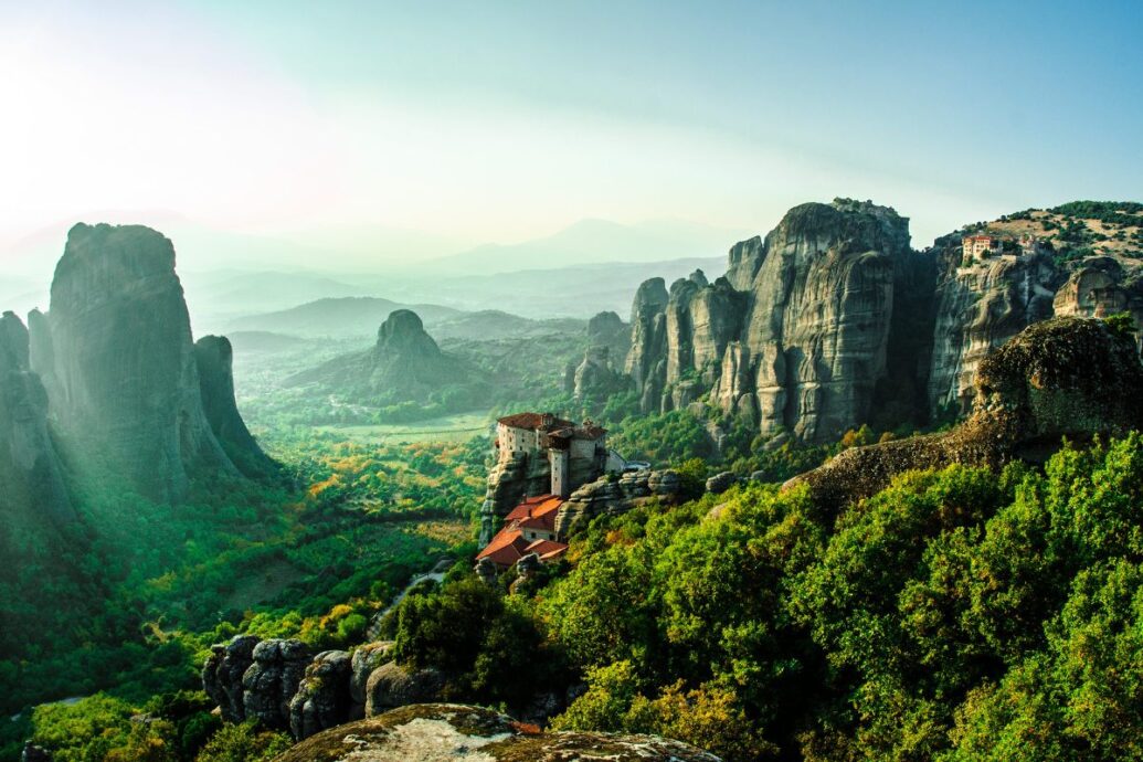 A scenic view of Meteora, Greece, showcasing towering rock formations with historic monasteries perched atop cliffs, surrounded by lush greenery and bathed in soft, natural light.